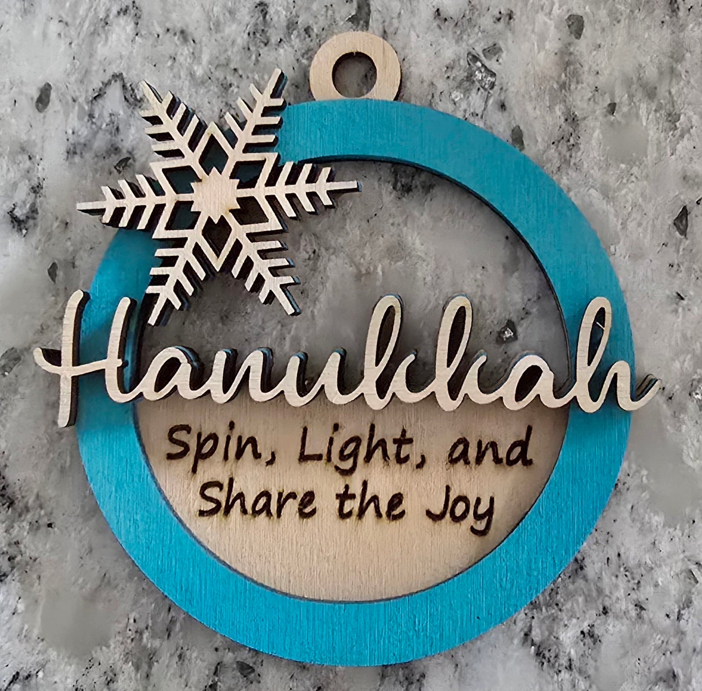 A circular wooden Hanukkah ornament with a blue background, featuring a silver snowflake design and the text 'HANUKKAH' in the center, with the phrases 'SPIN, LIGHT, AND SHARE THE JOY' below the snowflake.