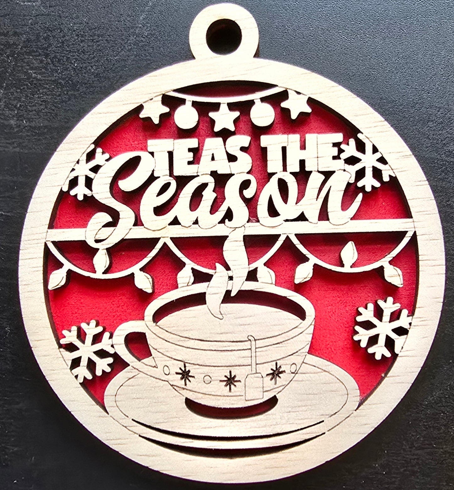A circular ornament with a red background, featuring the text 'TEAS THE SEASON' in white, surrounded by snowflake designs, a cup of tea, and a teapot.
