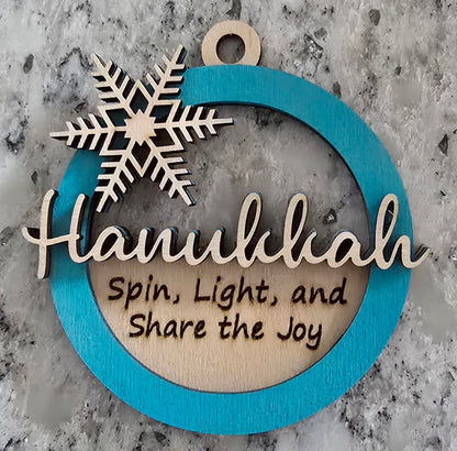 A circular wooden Hanukkah ornament with a blue background, featuring a silver snowflake design and the text 'HANUKKAH' in the center, with the phrases 'SPIN, LIGHT, AND SHARE THE JOY' below the snowflake.