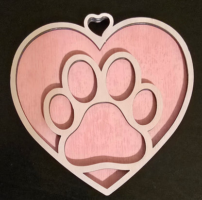 A wooden heart-shaped ornament with a paw print design cut out from the center, painted in shades of pink and white, with a small hook for hanging.