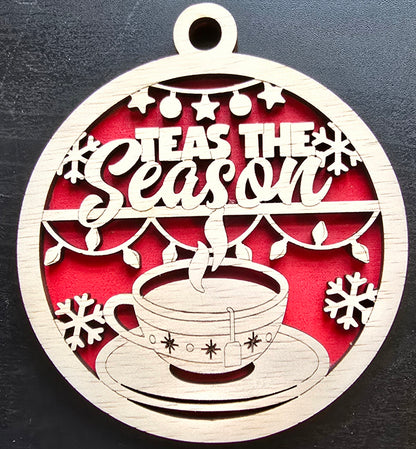 A circular ornament with a red background, featuring the text 'TEAS THE SEASON' in white, surrounded by snowflake designs, a cup of tea, and a teapot.